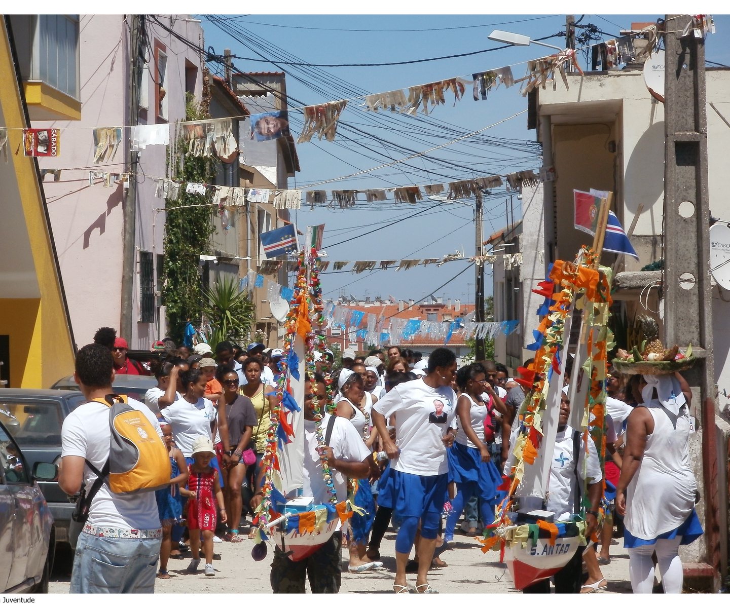 Desfile do Kola San Jon na Cova da Moura: homens carregam barcos decorados e mulheres de branco e azul dançam na rua
